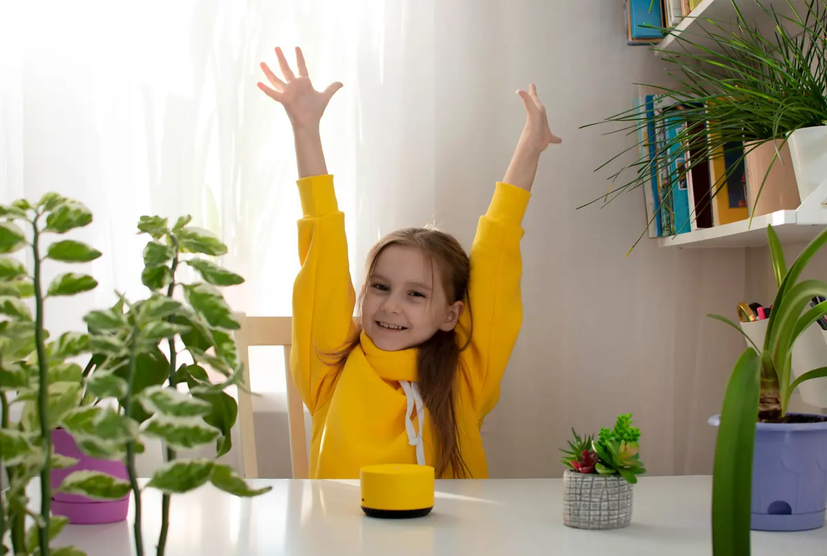 A girl at a table throws her arms up in celebration beside a small smart speaker.